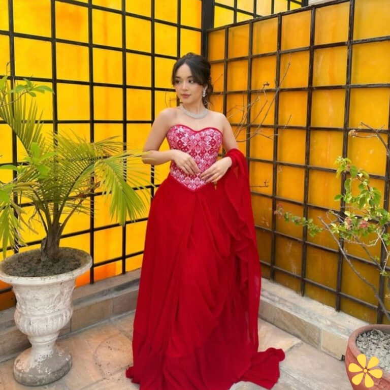 Woman in a stunning red gown stands by a vibrant yellow backdrop with plants.