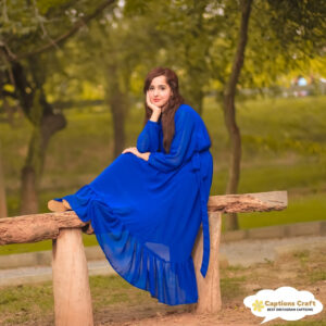 A woman in a flowing blue dress sits thoughtfully on a wooden log in a serene park.